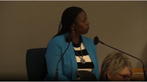 A woman in a striped shirt and blue jacket sits at a microphone in court