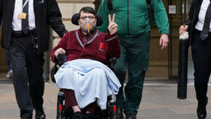 A man holds up two fingers in a peace sign as he sits in a wheelchair with a blanket over his lap
