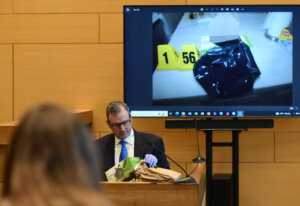 Man sits on witness stand with images of evidence on monitor behind him