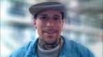 smiling tattooed gut with a blue shurt, brown beaded necklace and a cap.