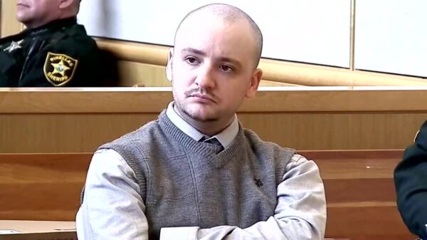 young bald man in court wears grey sweater vest over a shirt and tie